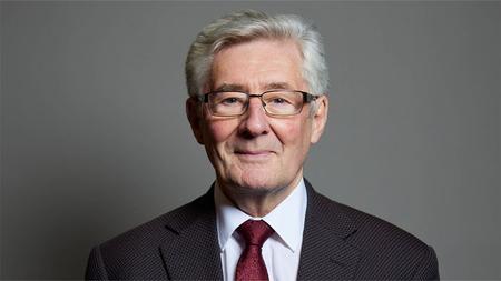 A portrait of Tony Lloyd MP, wearing a suit with a red tie and standing in front of a grey background.